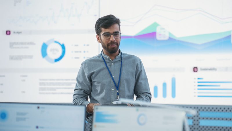 Ein Mann mit Brille und hellblauem Hemd steht vor einem großen Bildschirm, auf dem verschiedene Grafiken und Datenvisualisierungen in einer Büroumgebung angezeigt werden.