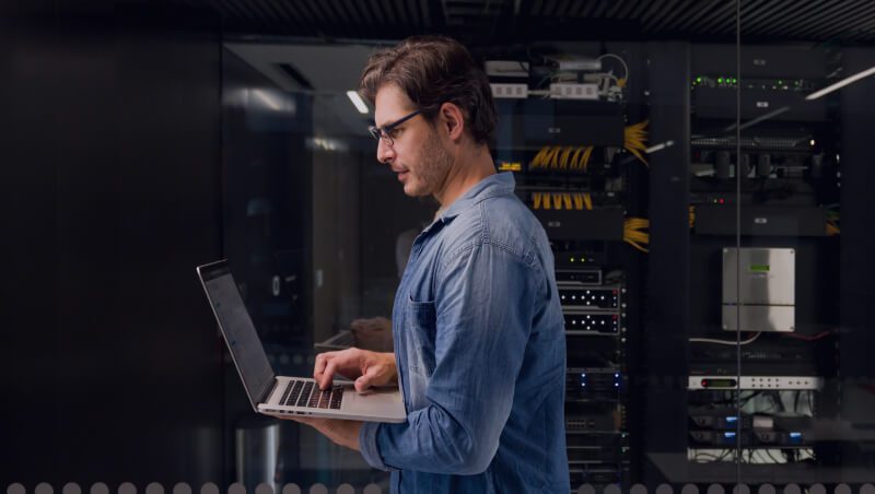 Ein Mann in einem blauen Hemd, der einen Laptop in einem Serverraum mit Reihen von Servern und Netzwerkgeräten im Hintergrund benutzt. Diese Umgebung spiegelt den Fokus auf die Operationalisierung von Sicherheit und Nonstop SecOps wider.