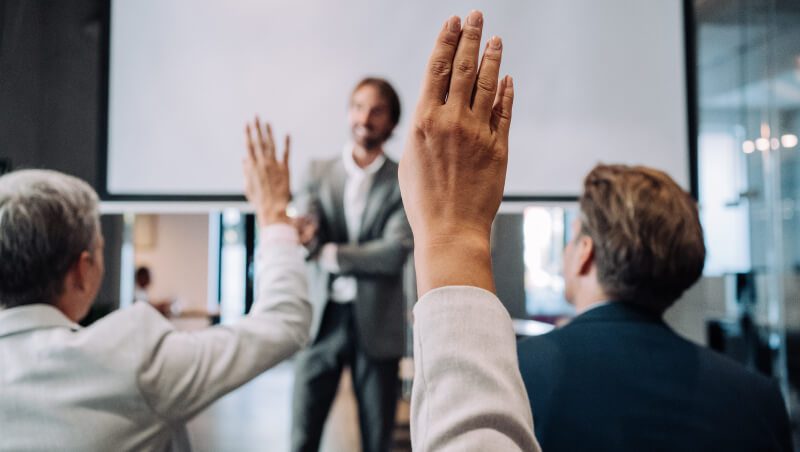 Eine Gruppe von drei Personen in Geschäftskleidung, von denen zwei ihre Hände heben, steht vor einem Bildschirm in einem modernen Besprechungsraum und blickt zu einem Redner.