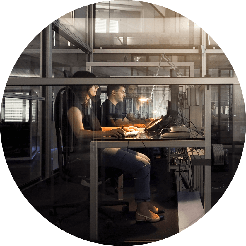 Zwei Personen arbeiten an Computern in einem modernen Büro mit Glastrennwänden. Eine Frau ist auf ihren Bildschirm konzentriert, während ein Mann neben ihr sitzt. Beide werden von warmem Licht beleuchtet.