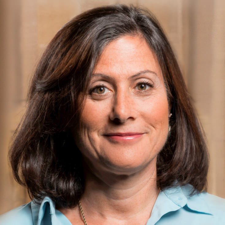 Gavriella Schuster trägt eine hellblaue Bluse und blickt mit neutralem Gesichtsausdruck direkt in die Kamera. Der Hintergrund ist weich und unscharf, was auf eine professionelle Umgebung hindeutet.