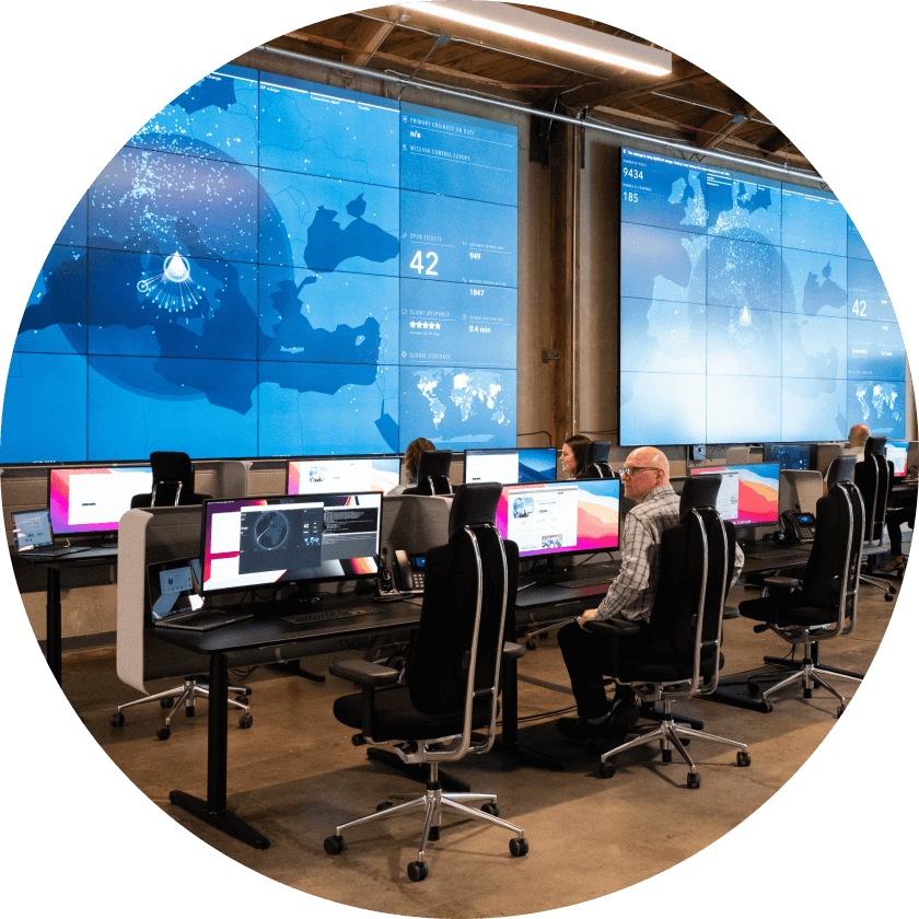 Ein modernes Büro mit mehreren Arbeitsplätzen und großen digitalen Bildschirmen, auf denen Datenanalysen, Karten und Statistiken angezeigt werden. Mehrere Personen sitzen vor ihren Monitoren und sind konzentriert bei der Arbeit.