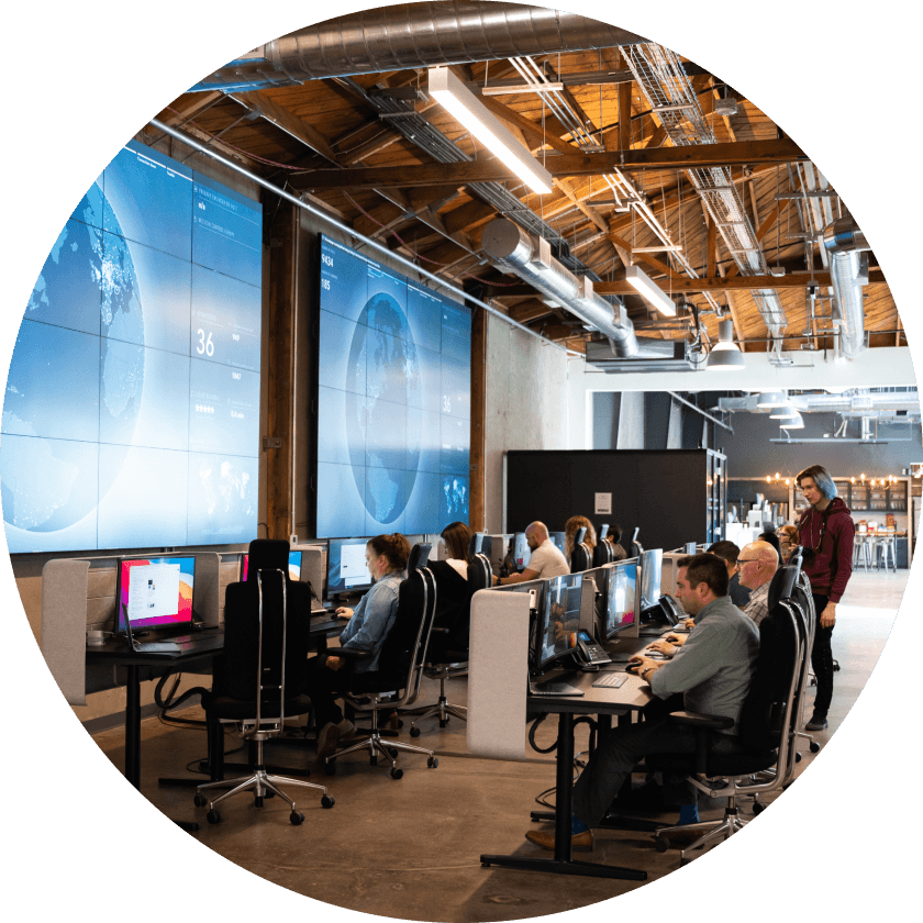 Ein Team von Fachleuten arbeitet an Computern in einem modernen Büro, im Hintergrund sind große Bildschirme zu sehen, auf denen Daten und Karten angezeigt werden. Der Arbeitsbereich ist mit einer Mischung aus Schreibtischen und Stühlen ausgestattet.