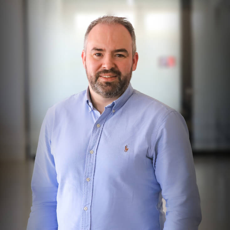 Craig Jones trägt ein hellblaues Hemd mit Knöpfen und steht lächelnd in einer modernen Büroumgebung.