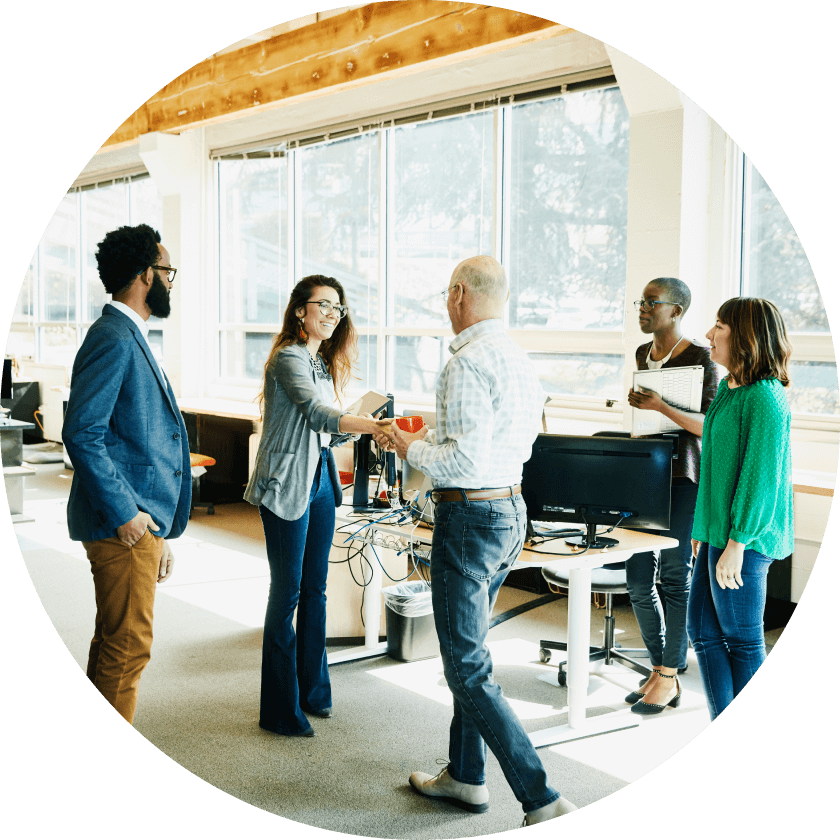 Eine Gruppe von fünf Fachleuten in einer Büroumgebung. Eine Frau gibt einem Mann die Hand, während drei andere zusehen. Das Büro hat große Fenster, Schreibtische und Computer sind zu sehen.