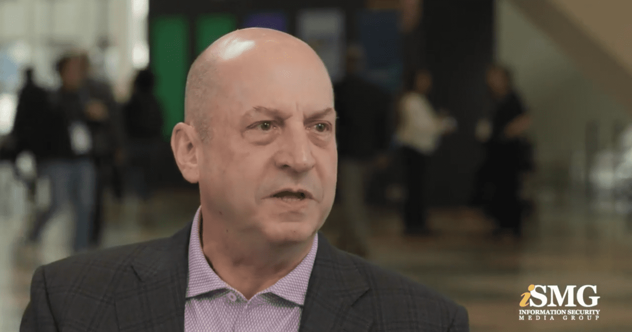 Ein Mann mit kurzen, hellen Haaren in einem dunklen Blazer und einem lila Hemd spricht während eines Interviews auf einer Konferenz. Im Hintergrund sind unscharfe Personen und Konferenzschilder zu sehen.