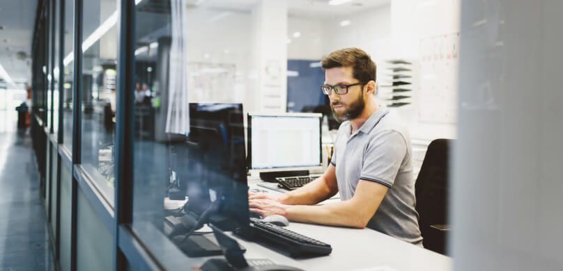 Ein Mann sitzt an einem Schreibtisch in einem Büro und arbeitet am Computer. Er trägt eine Brille und ein graues Polohemd mit kurzen Ärmeln. Auf dem Schreibtisch liegen Unterlagen und ein zweiter Monitor.