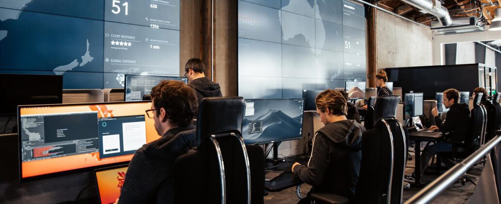 Ein modernes Büro, in dem mehrere Personen an Computertischen arbeiten und sich auf Bildschirme konzentrieren, auf denen Daten und Analysen angezeigt werden, während im Hintergrund große Videowände Karten und Statistiken zeigen.