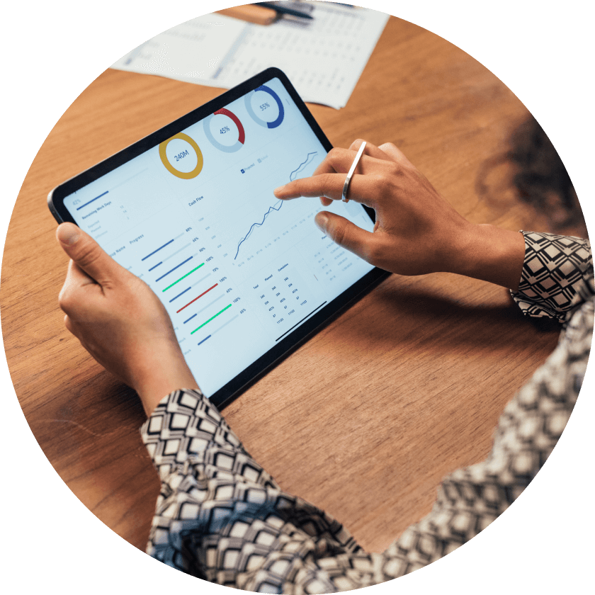 Eine Person analysiert Daten auf einem Tablet und zeigt Diagramme und Grafiken auf einem Holztisch an. Die Person trägt ein gemustertes Hemd und zeigt mit einem Stylus auf den Bildschirm.