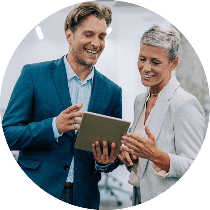 Ein Mann und eine Frau in Business-Kleidung unterhalten sich lächelnd, während sie in einer modernen Büroumgebung auf ein Tablet schauen.