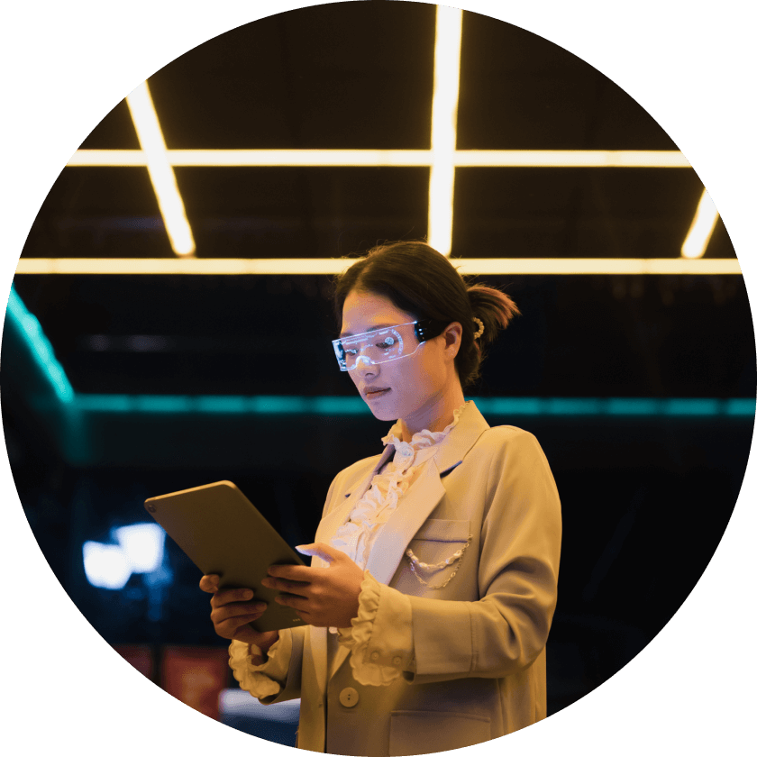 Eine Frau mit einer klaren Schutzbrille hält ein Tablet in der Hand, während sie unter einer hellen, modernen Beleuchtung steht. Sie ist mit einem hellen Blazer bekleidet und konzentriert sich auf das Gerät.