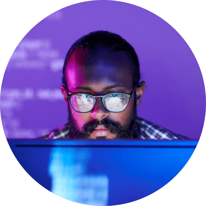 Eine Person mit Brille und Bart konzentriert sich auf einen Computerbildschirm, auf dem ein Code angezeigt wird, mit einem lila Hintergrund, der von verschiedenen Farben beleuchtet wird.