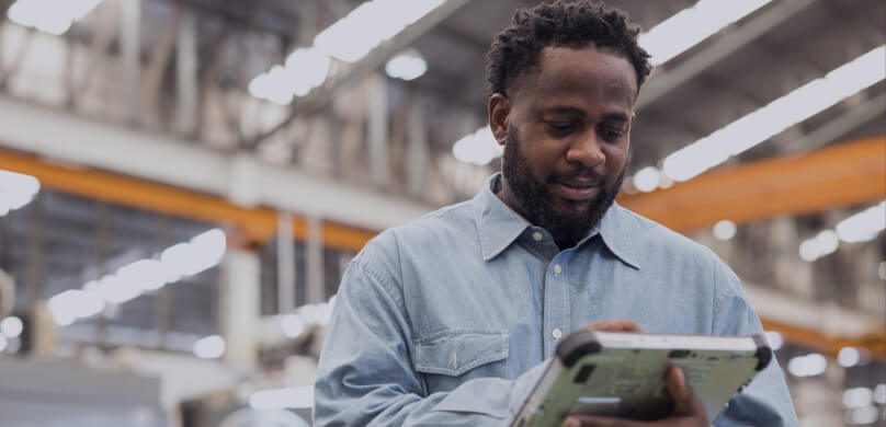 Ein Mann in einem blauen Hemd benutzt ein Tablet, während er in einem großen Industriegebäude mit hohen Decken und Deckenleuchten steht.