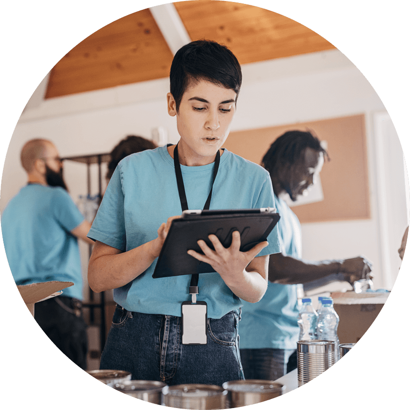 Eine junge Frau in einem blauen Hemd hält ein Klemmbrett in der Hand und konzentriert sich auf ihre Aufgabe. Im Hintergrund sortieren andere Freiwillige Gegenstände auf Tischen und arbeiten in einem hellen Raum.