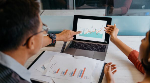 Zwei Fachleute besprechen während einer Besprechung Daten. Eine Person zeigt auf einen Laptop-Bildschirm, auf dem Grafiken angezeigt werden, während die andere Person auf den Bildschirm mit ausgedruckten Berichten auf dem Tisch deutet.