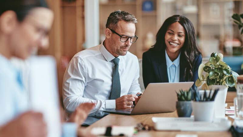 Ein Mann und eine Frau unterhalten sich, während sie in einem modernen Büro auf einen Laptop schauen. Der Mann trägt eine Brille und eine Krawatte, während die Frau in einem professionellen Outfit lächelt. Andere Kollegen sind im Hintergrund verschwommen zu sehen.