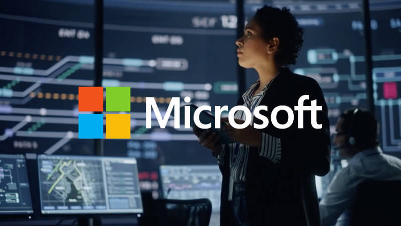 Eine Frau steht in einem Datenanalyse-Raum und betrachtet digitale Bildschirme, auf denen technische Grafiken und Informationen angezeigt werden. Im Hintergrund sitzt eine Person an einem Schreibtisch. Das Microsoft-Logo ist deutlich zu sehen.