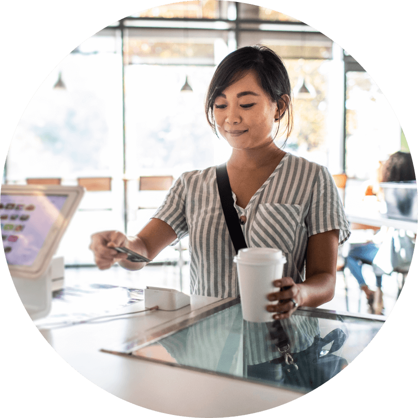 Eine Frau in einem gestreiften Hemd steht an einer Café-Theke und bezahlt mit einer Karte, während sie eine Kaffeetasse in der Hand hält. Der Innenraum verfügt über große Fenster und sanfte Beleuchtung.