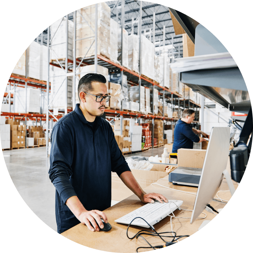 Ein Mann mit Brille arbeitet an einem Computer in einem Lagerhaus, im Hintergrund sind Regale mit Kartons zu sehen. Im Hintergrund ist ein weiterer Arbeiter zu sehen, der Gegenstände sortiert.