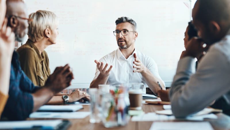 Eine Gruppe von Fachleuten, die an einem Holztisch eine Besprechung abhalten, wobei ein Mann spricht und gestikuliert, während andere aufmerksam zuhören. Auf dem Tisch sind Papiere und Tassen zu sehen.