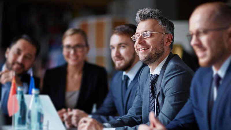 Eine Gruppe von fünf Fachleuten sitzt um einen Tisch herum und diskutiert. Sie tragen Geschäftskleidung, in der Nähe sind Flaggen von Deutschland und Frankreich zu sehen. Der Rahmen scheint eine Unternehmenskonferenz oder ein Meeting zu sein.