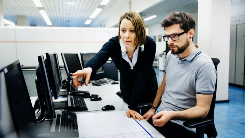 Eine Frau zeigt auf einen Computerbildschirm, während sie einem Mann, der neben ihr in einem Büro sitzt, etwas erklärt. Im Hintergrund sind mehrere Computer zu sehen.