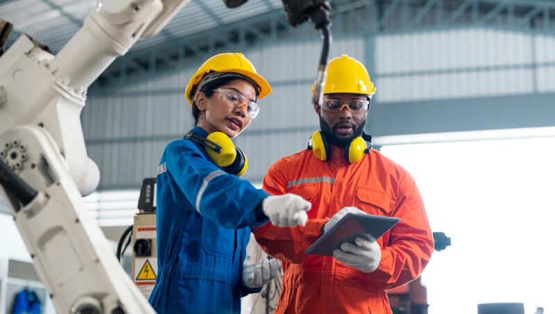 Zwei Arbeiter besprechen technische Details in einer Fabrikumgebung. Der eine trägt eine blaue Arbeitsuniform und einen gelben Helm, der andere einen orangefarbenen Overall und einen gelben Helm. Im Hintergrund ist ein Roboterarm zu sehen.