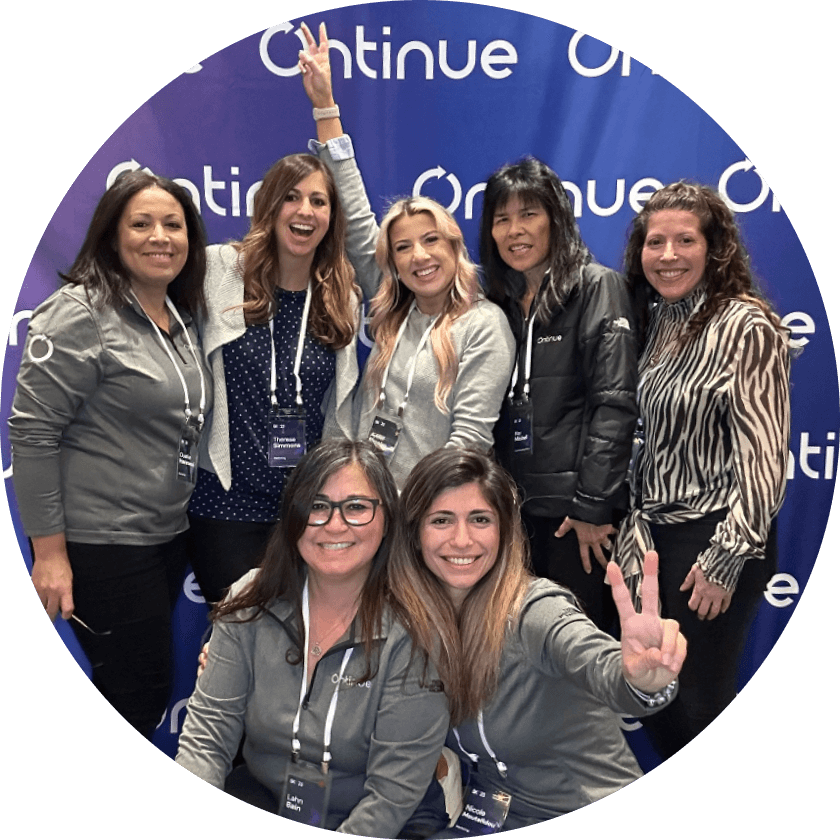 Eine Gruppe von sieben Frauen posiert für ein Foto vor einem blauen Hintergrund mit dem Wort 'Continue' in Weiß. Die Frauen tragen passende graue Jacken und lächeln, wobei eine Frau ein Friedenszeichen macht.