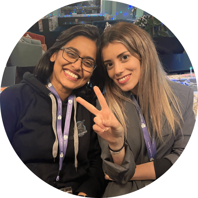 Zwei Frauen sitzen lächelnd beieinander, eine macht mit ihrer Hand ein Friedenszeichen. Beide tragen Schlüsselbänder und Freizeitkleidung vor einem festlichen Hintergrund mit sanfter Beleuchtung.