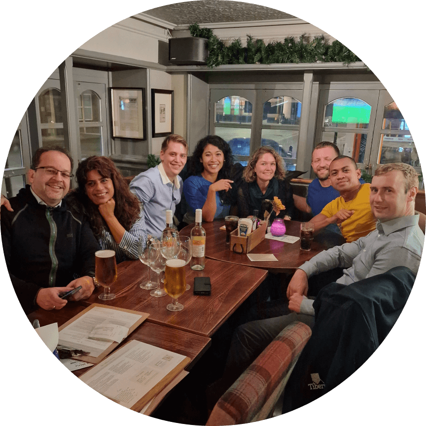 Eine Gruppe von acht Personen sitzt an einem Holztisch in einer Kneipe und genießt Getränke und Essen. Die Umgebung besteht aus Biergläsern, Flaschen und einer Speisekarte. Die Atmosphäre wirkt entspannt und gesellig.