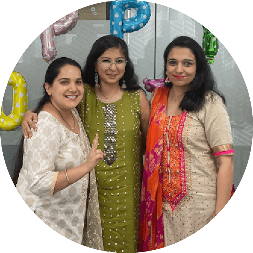 Drei Frauen in traditioneller Kleidung posieren gemeinsam auf einer Feier. Sie lächeln und stehen vor farbenfrohen Ballondekorationen mit dem Schriftzug 'UP'. Im Hintergrund ist eine moderne Büroeinrichtung zu sehen.