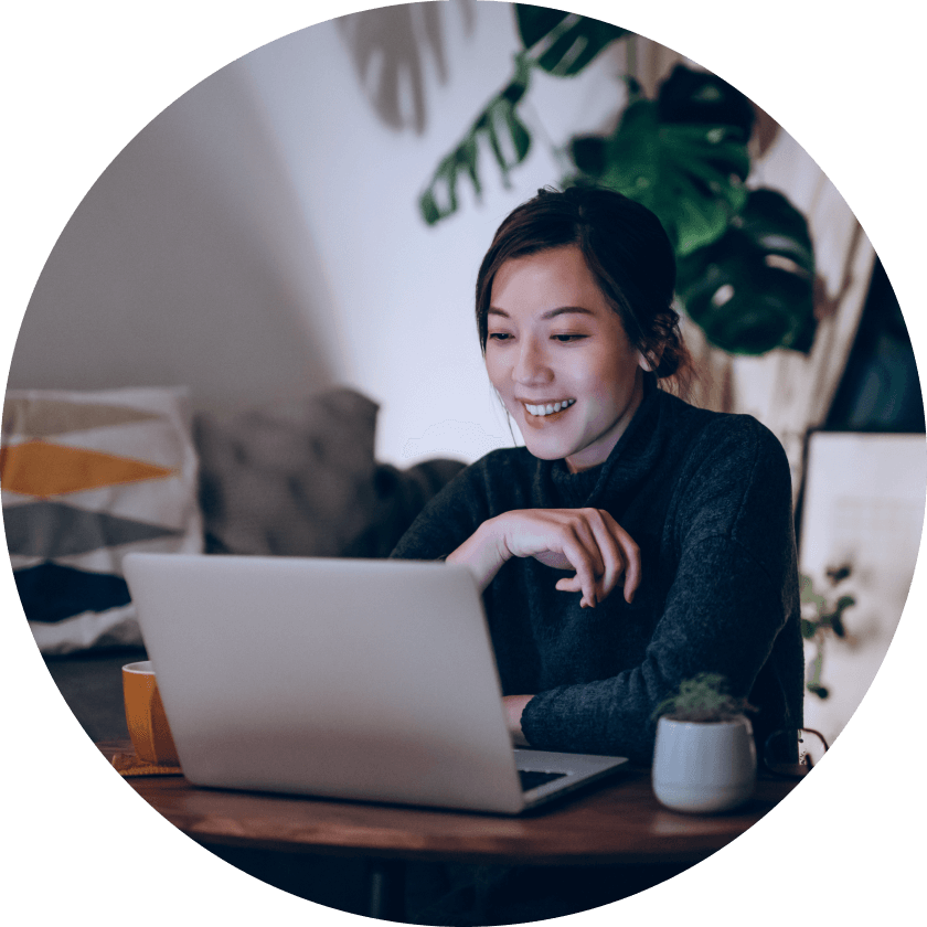 Eine Frau sitzt an einem Holztisch mit einem Laptop vor sich und lächelt bei der Arbeit. Sie befindet sich in einem gemütlichen Raum mit Pflanzen und einer Couch im Hintergrund.