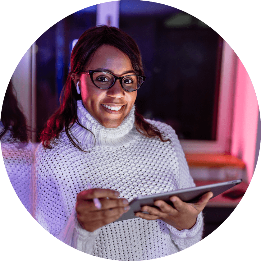 Eine Frau, die einen gemütlichen weißen Pullover und eine Brille trägt, lächelt, während sie in einem schwach beleuchteten Raum ein Tablet in der Hand hält, mit bunten Lichtreflexen im Hintergrund.