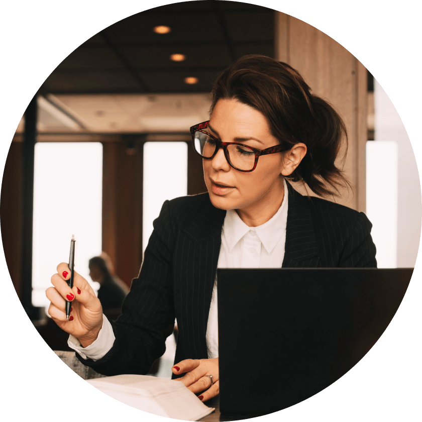 Eine Frau in einem Nadelstreifenanzug und mit Brille sitzt an einem Tisch mit einem Laptop. Sie hält einen Stift in der Hand und scheint mit der Arbeit beschäftigt zu sein, vor sich liegen Notizen und Dokumente.