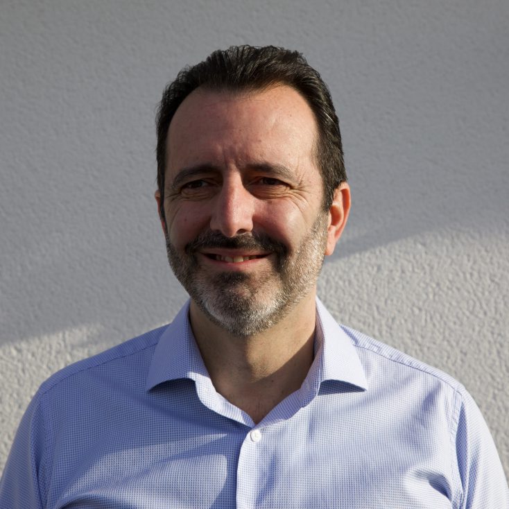 Roberto Veiga, lächelnd, in einem hellblauen Hemd, vor einer weißen Wand stehend.