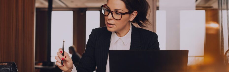 Eine Frau mit Brille und Blazer sitzt an einem Tisch und schreibt mit einem Stift, während sie auf einen Laptop schaut. Die Umgebung scheint ein modernes Büro oder Café zu sein.