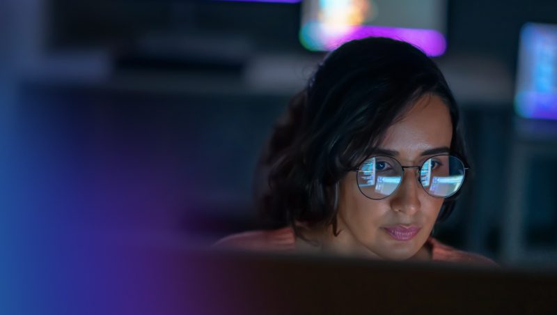 Eine Frau mit Brille arbeitet an einem Computer in einer schwach beleuchteten Umgebung, im Hintergrund sind bunte Bildschirme zu sehen. Der Fokus liegt auf ihrem konzentrierten Gesichtsausdruck, während sie Daten oder Informationen prüft. NIS2 erklärt.