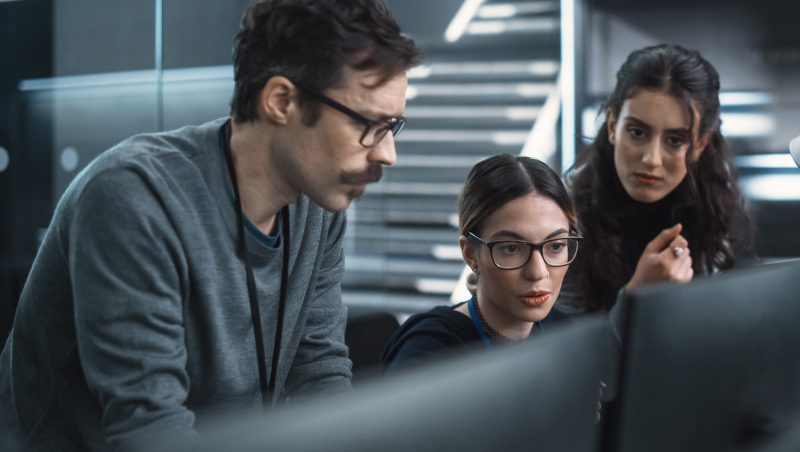 Drei Kollegen diskutieren in einem Hightech-Büro über Datenanalyse. Zwei Frauen und ein Mann konzentrieren sich auf einen Computerbildschirm und betonen die Zusammenarbeit im Hinblick auf Sicherheitsbedrohungen im Jahr 2023.