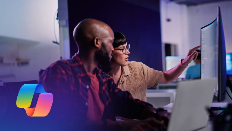 Ein Mann und eine Frau arbeiten an einem Computerarbeitsplatz zusammen und besprechen ihre Arbeit auf dem Bildschirm. Die Umgebung ist modern und gut beleuchtet, auf den Bildschirmen werden Programme oder digitale Inhalte angezeigt. Das Bild enthält ein Logo in der unteren linken Ecke, das auf Microsoft Copilot für Sicherheit verweist.