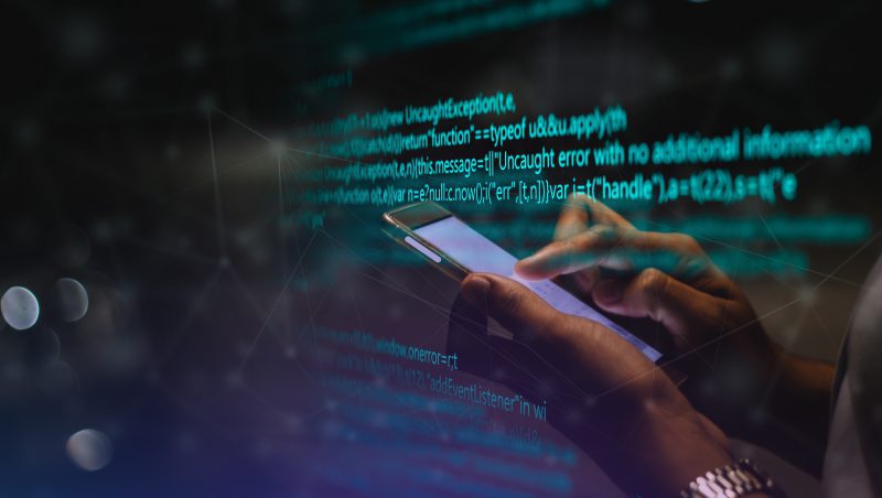 Eine Person hält ein Smartphone in der Hand, während sie auf einem Bildschirm im Hintergrund Programmiercode sieht. Der Code enthält Fehlermeldungen und Programmiersyntax und schafft eine technisch orientierte Atmosphäre, die für digitale Sicherheit und Fehlerbehebung relevant ist, einschließlich PlugX Utilising Steam.