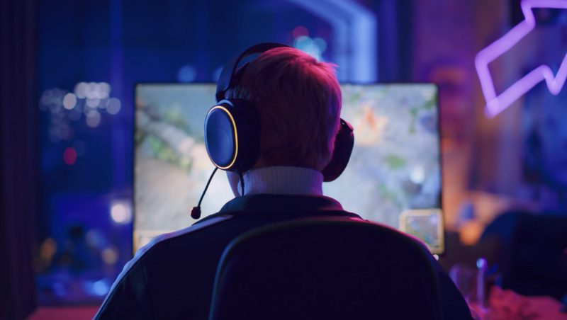 Eine Person mit kurzen Haaren, die ein Headset trägt, sitzt vor einem Computermonitor, auf dem ein Videospiel angezeigt wird. Der Raum ist mit bunten LED-Lichtern beleuchtet, die eine Spielatmosphäre schaffen.
