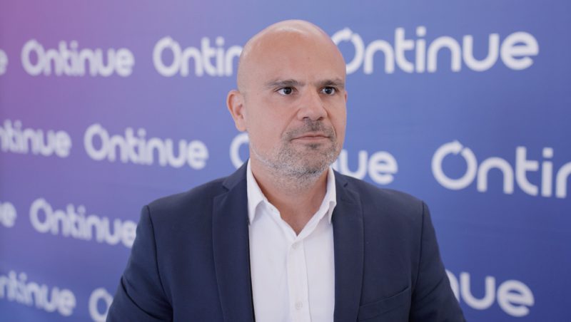 Ein Mann mit Glatze und hellem Bart, bekleidet mit einem dunklen Anzug und einem weißen Hemd, steht vor einem blauen Hintergrund mit dem Logo 'Ontinue' in weißer Schrift.