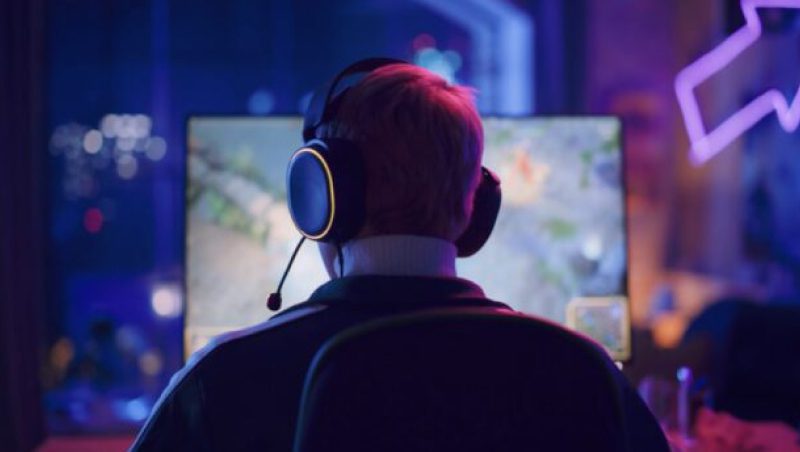 Eine Person sitzt in einem Gaming-Stuhl, trägt ein Headset und konzentriert sich auf einen Computerbildschirm, auf dem ein Videospiel angezeigt wird. Die Szene wird durch sanftes, farbenfrohes Licht aus dem Fenster und die Raumdekoration beleuchtet.