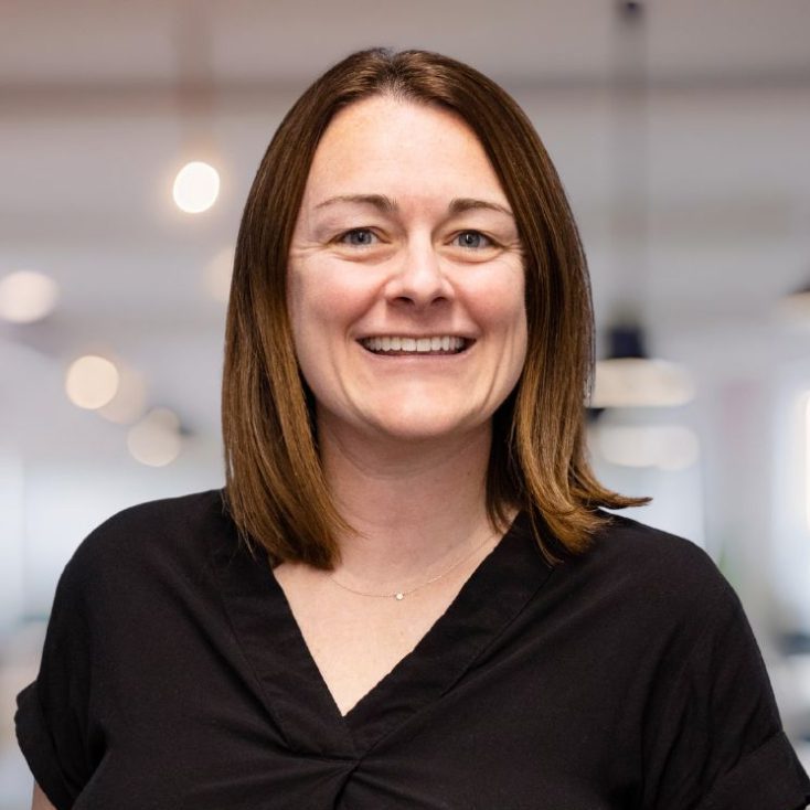 Kate Montgomery, bekleidet mit einer schwarzen Bluse, lächelt in eine Kamera in einer modernen Büroumgebung mit unscharfen Lichtern im Hintergrund.