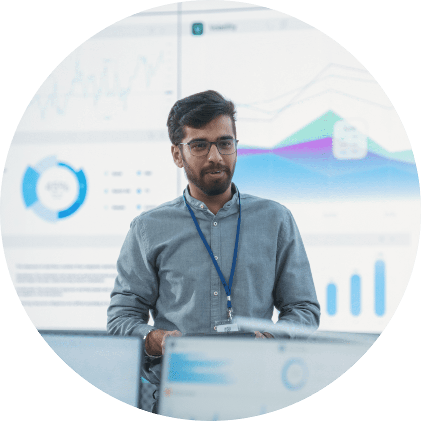 Ein Mann in einem hellblauen Hemd und mit Brille steht vor einem großen Bildschirm, auf dem Grafiken und Datenvisualisierungen in einer modernen Büroumgebung angezeigt werden.