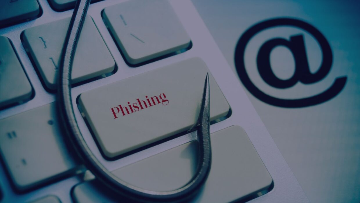 Nahaufnahme einer Tastatur, auf der ein Angelhaken auf der Taste 'Phishing' liegt, neben einem '@'-Symbol im Hintergrund. Dieses Bild veranschaulicht das Konzept von Salty2FA Multi-Stage Phishing.