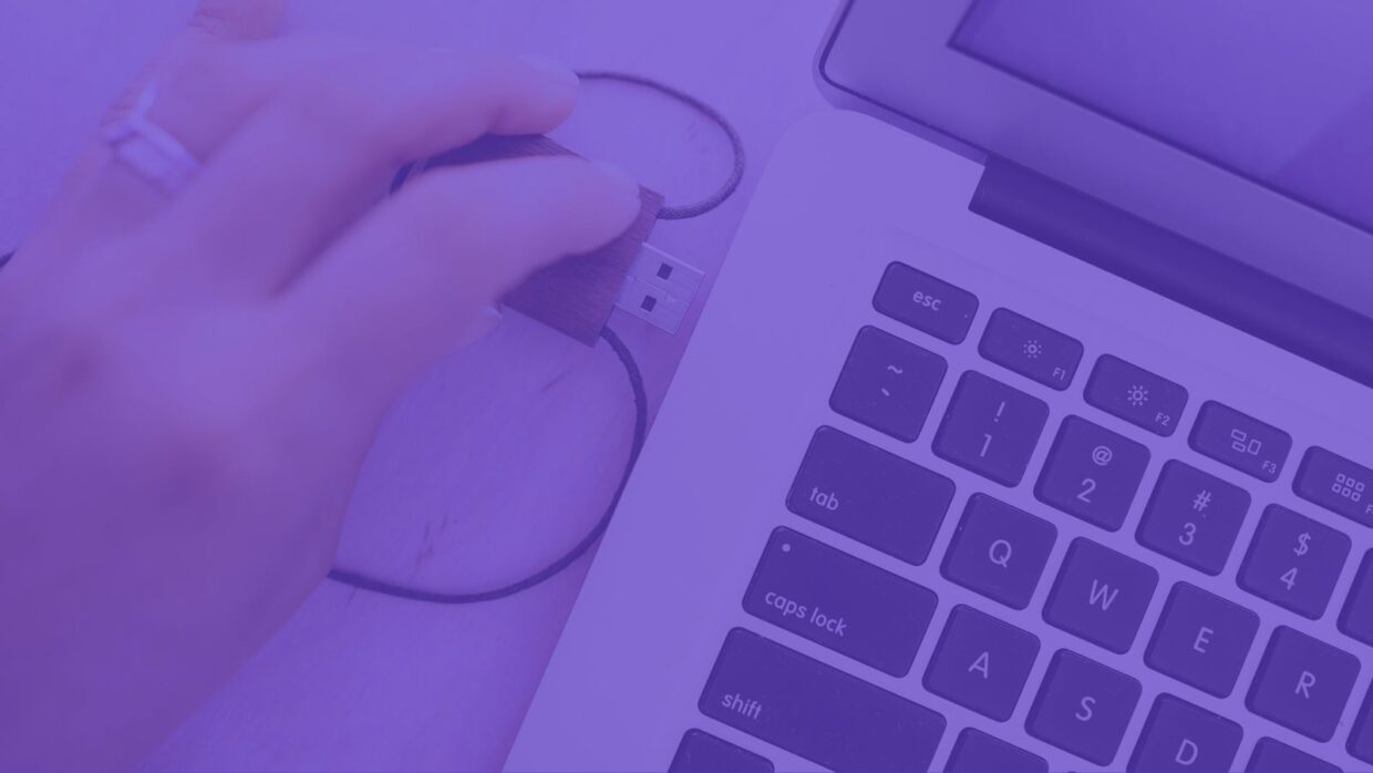 Eine Hand hält einen USB-Stick über den USB-Anschluss eines Laptops, wobei die Tastatur des Laptops zu sehen ist. Das Bild ist violett getönt, um die Aktion des Anschließens der USB-Malware an das Gerät zu betonen.