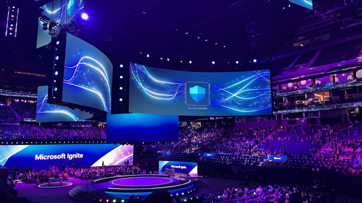 Ein großes Auditorium voller Zuschauer, die an der Microsoft Ignite-Veranstaltung teilnehmen. Auf der Bühne befinden sich mehrere große Bildschirme, auf denen lebendige blaue Grafiken mit dem Microsoft Ignite-Logo im Mittelpunkt zu sehen sind.