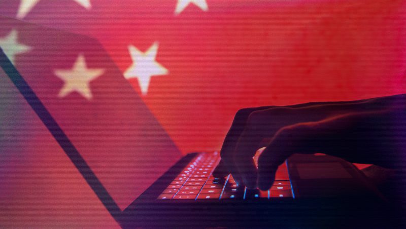Eine silhouettierte Hand, die auf einem Laptop mit beleuchteten Tasten tippt, vor dem Hintergrund einer roten Fläche mit weißen Sternen, die an die chinesische Flagge erinnert.