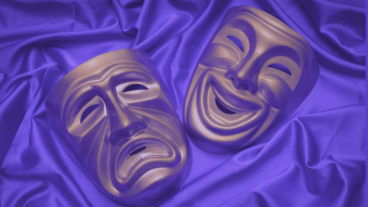 Zwei Theatermasken auf einem violetten Satinstoff: eine Maske stellt die Komödie mit einem lächelnden Ausdruck dar, die andere die Tragödie mit einem stirnrunzelnden Ausdruck.